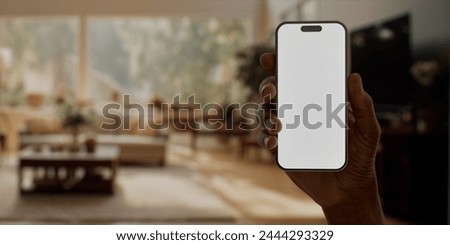 Black African-American man using smartphone with a blank white screen in a sunlit living room, modern lifestyle and connectivity apps mockup. Vertical phone orientation