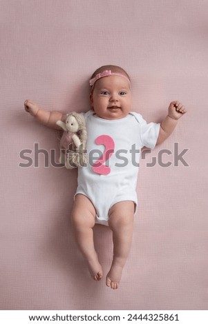 girl baby infant with a bow lies on the bed in a white bodysuit with a doll toy next to it, the child has a pink number 2 months on a pink background.