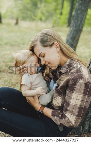 A cute mother is sitting in the park on the grass with a small child in her arms. A walk with a small child in nature.