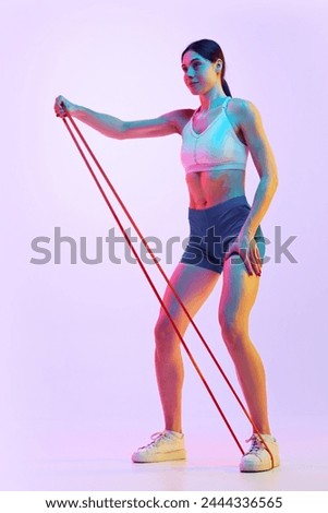 Side view of focused, brunette woman exercising with resistance band in neon light against gradient studio background. Concept of sport and recreation, movement, self care, action, energy. Ad