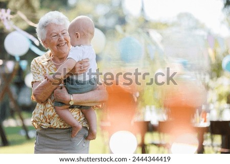 Great-grandmother holding little baby in her arms. Family summer garden party. Royalty-Free Stock Photo #2444344419