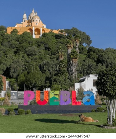 A colorful PUEBLA sign in front of a lush green hill. The hill is topped by a church with a yellow facade and a large dome. In the foreground, a brown dog lounges in the grass.