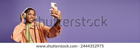 Cheerful african american woman in 20s taking selfie with her headphones on lilac backdrop, banner