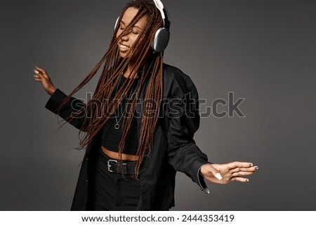 joyful and young African American woman in 20s with headphones dancing to music on grey backdrop Royalty-Free Stock Photo #2444353419