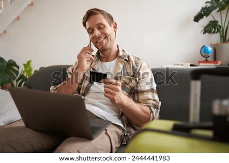 Portrait of smiling guy, tourist booking hotel, confirming purchase over the phone, holding credit card and laptop. Royalty-Free Stock Photo #2444441983