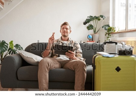 Portrait of man waiting for his holiday tour confirmation, cross fingers, hopes for something, holds tablet, has suitcase packed and ready for holiday. Royalty-Free Stock Photo #2444442039