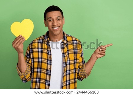 Portrait of positive guy beaming smile hold paper heart symbol card direct finger empty space isolated on green color background