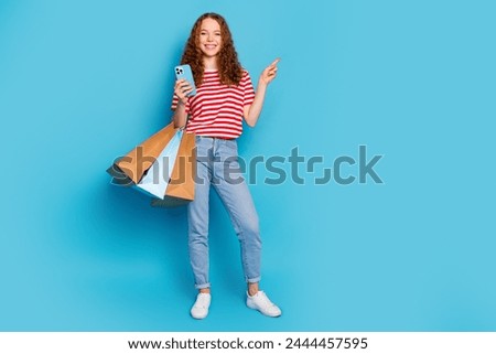 Full length photo of pleasant woman hold shopping bags smartphone indicating at offer empty space isolated on blue color background
