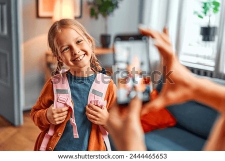 Picture from childhood. Delighted kid looking at cell phone camera with toothy smile and holding handles of pink schoolbag. Happy caucasian girl standing at home while mother taking picture.