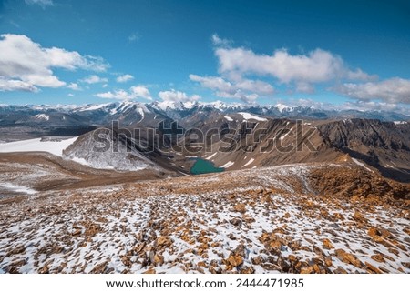 Sunny top view to most beautiful alpine lake between stony hill in freshly fallen snow and sharp rocky ridge in sunlight. Large colorful mountain range with snow-white peaks in far away in low clouds. Royalty-Free Stock Photo #2444471985