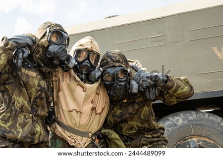 Three soldiers wearing gas masks are posing for a picture. Scene is serious and tense, as the soldiers are wearing protective gear and appear to be in a dangerous situation