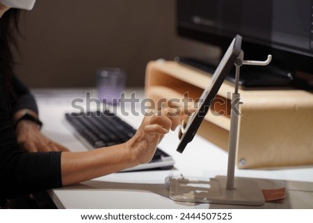 Close up image, Selective focus unrecognizable woman in mask use finger touch on screen of digital tablet on stand at workplace