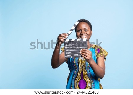 African american girl posing with clapperboard in front of camera, smiling and using film slate in the post production movie industry. Female model working in filmography or videography.