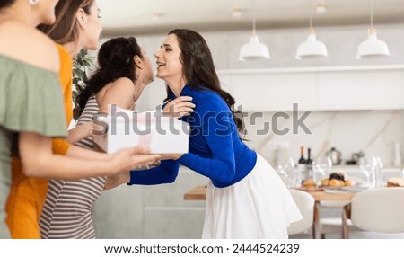 Hospitable hostess in vibrant festive attire warmly greeting and kissing female friends arriving for home get-together in cozy atmosphere with set table Royalty-Free Stock Photo #2444524239