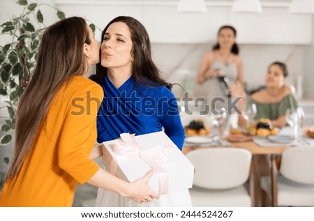 Hospitable hostess in vibrant festive attire warmly greeting and kissing female friends arriving for home get-together in cozy atmosphere with set table Royalty-Free Stock Photo #2444524267