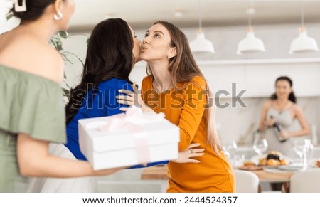 Hospitable hostess warmly greeting and kissing female friends arriving for home get-together with food and drinks on table in background.. Royalty-Free Stock Photo #2444524357