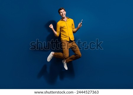 Full size photo of handsome young man jumping hold device raise fist dressed stylish yellow outfit isolated on dark blue color background