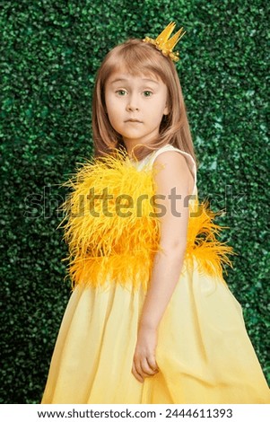 Portrait of a cute little princess girl standing in a festive yellow dress and a golden crown by a green boxwood background. Kid’s fashion. Birthday. March 8, Women's Day.