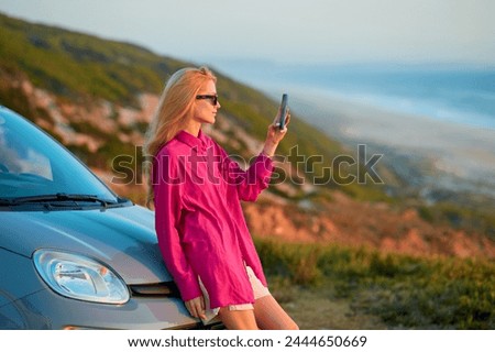 A woman takes a photo of a sunset by the sea leaning on a car.