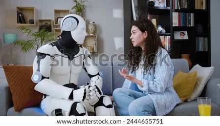 Side view of serious young woman sitting with humanoid robot on comfortable sofa in living room. Automated bionic cyborg listening carefully female's story indoors. Royalty-Free Stock Photo #2444655751