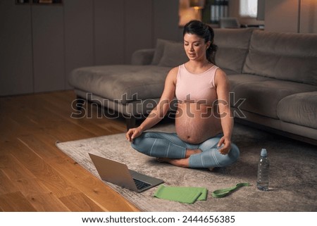A beautiful young pregnant woman following a video tutorial on her laptop while doing breathing exercises and sitting on the floor in the living room.