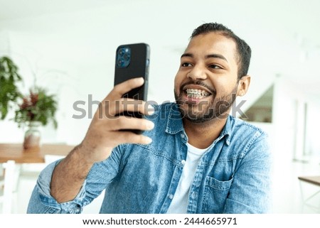 cheerful african american man with braces uses smartphone and takes pictures in white room, man in denim shirt holds mobile phone and photographing