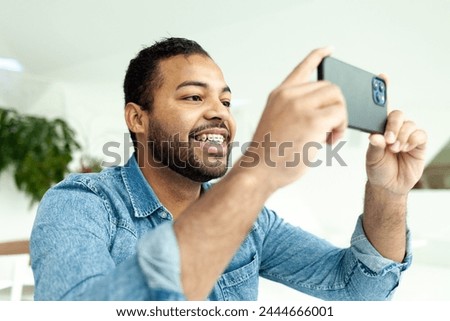 cheerful african american man with braces uses a smartphone and takes pictures in a white room, a man in a denim shirt holds a mobile phone horizontally and watches a video