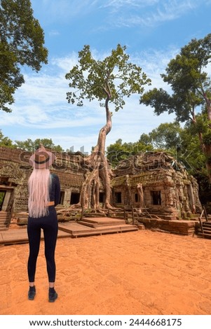 A blonde woman wearing black tights and a beige hat is standing -Ta Prohm temple ruins hidden in jungles at Angkor Wat - Wall carving with woman famous Angkor Wat complex, Siem Reap, Cambodia