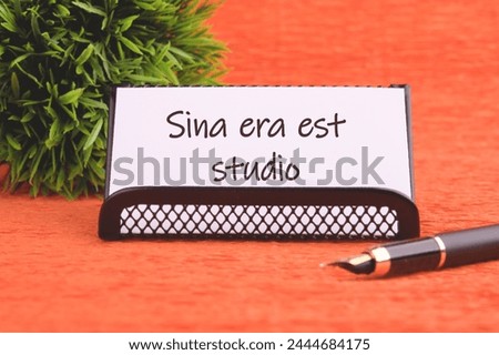 Sina era est studio It means Without anger and addiction on a white business card on an orange background, next to a fountain pen and a plant in the background