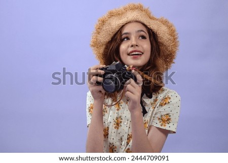 Smiling pre-teen girl wears straw hat taking picture on retro camera in studio on violet background