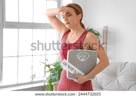 Sporty mature woman holding paper with word MENOPAUSE and weight scales at home Royalty-Free Stock Photo #2444732025