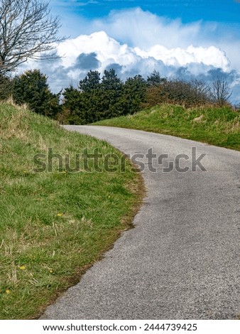 A winding road bending uphill to the left with grass verges, trees and clouds at Curracloe, Wexford, Ireland.  Royalty-Free Stock Photo #2444739425