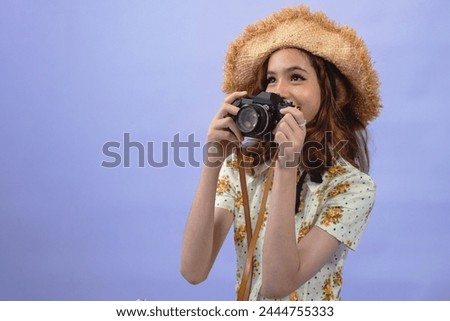 Smiling pre-teen girl wears straw hat taking picture on retro camera in studio on violet background