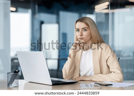 Thoughtful female professional contemplating while working on a laptop in a contemporary office setting, looking at camera. Royalty-Free Stock Photo #2444833859