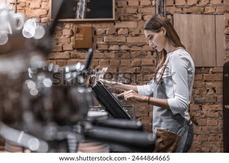 Young caucasian woman in apron working with cash box in coffee shop behind the counter. Female worker, barista, waitress, manager, administrator checking bill order on digital cash register. Royalty-Free Stock Photo #2444864665