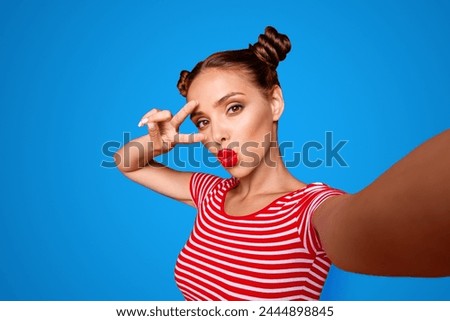Pretty cheerful girl with pouted lips shooting selfie and holding two fingers near eyes showing v-sign isolated on red background