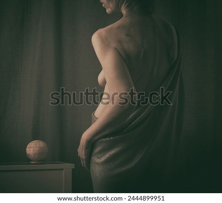back turned woman covering herself with a tunic in the style of a Greek statue recreating the art of baroque painters with chiaroscuro lighting in an interior with a piece of furniture and a bone ball Royalty-Free Stock Photo #2444899951