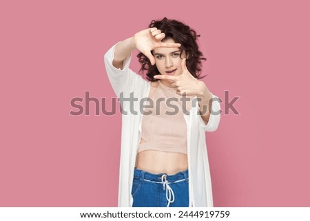 Portrait of concentrated attractive woman with curly hair wearing casual style outfit looking at camera through frame from her fingers. Indoor studio shot isolated on pink background.
