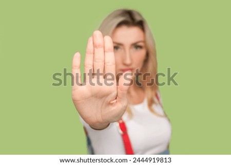 Portrait of serious strict adult blond woman showing stop sign, making forbidden gesture, looking at camera, wearing denim overalls. Indoor studio shot isolated on light green background