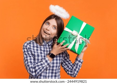 Portrait of cute curious woman with brown hair and nimb over head shaking present box thinking what inside, wearing checkered shirt. Indoor studio shot isolated on orange background