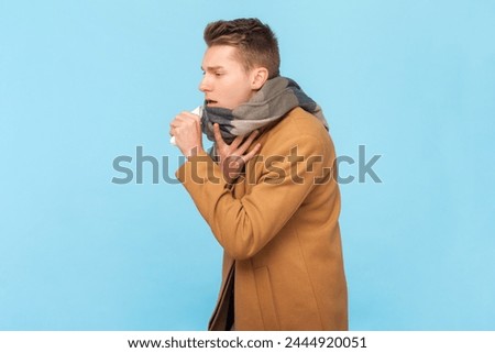 Portrait of sick ill young man wearing white warm jacket and scarf standing coughing, having flu symptoms, grippe, high temperature. Indoor studio shot isolated on blue background Royalty-Free Stock Photo #2444920051