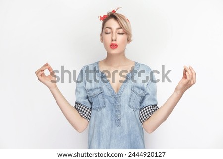 Portrait of relaxed calm blonde woman wearing blue denim shirt and red headband standing practicing yoga, meditating, keeps eyes closed. Indoor studio shot isolated on gray background.