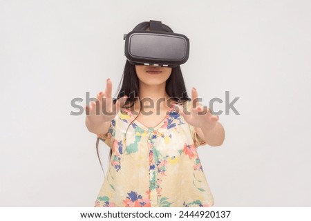 Portrait of playful woman with long brunette hair with vr headset playing game standing with outstretched hands. Indoor studio shot isolated on pink background. Royalty-Free Stock Photo #2444920137