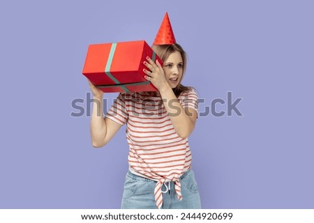 Portrait of curious blond woman wearing striped T-shirt and party cone shaking present box, thinking what inside, looking away with smile. Indoor studio shot isolated on purple background.