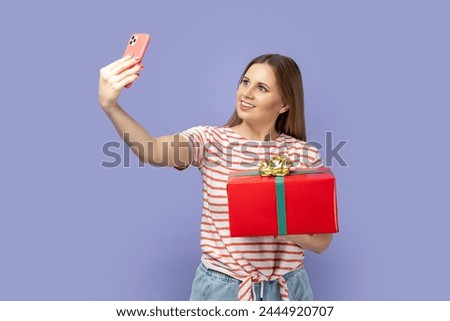 Portrait of optimistic blond woman blogger wearing striped T-shirt holding red present box and making selfie or broadcasting livestream. Indoor studio shot isolated on purple background.