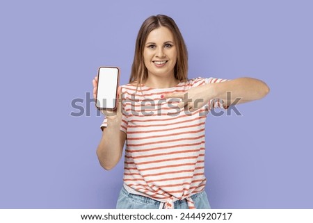 Portrait of friendly happy blond woman wearing striped T-shirt holding pointing at mobile phone with white empty display with advertising area. Indoor studio shot isolated on purple background.