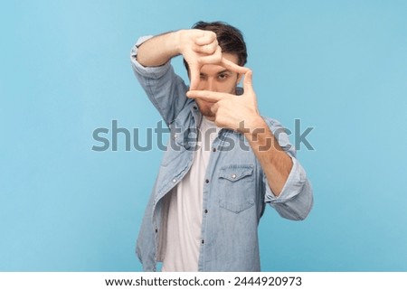 Portrait of focused unshaven attractive man photographer making frame with both hands, having satisfied facial expression, wearing denim shirt. Indoor shot isolated on blue background.