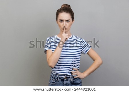 Portrait of serious woman wearing striped T-shirt standing holding finger on lips, making hush silence gesture, asking to keep secret. Indoor studio shot isolated on gray background.