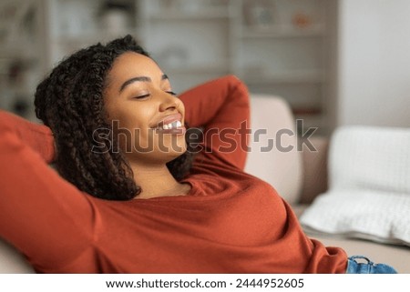 Content young black woman with a beaming smile reclines on a couch, her hands behind her head, eyes closed in serene repose, african american female resting at home Royalty-Free Stock Photo #2444952605