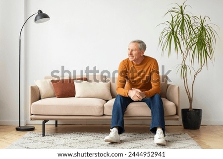 Senior man seated comfortably on a couch amidst warm-toned pillows, with a potted plant in the background Royalty-Free Stock Photo #2444952941